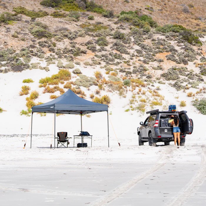 A woman works in her overland truck while her shade tent is secured into the sand with Tiegear equipment.